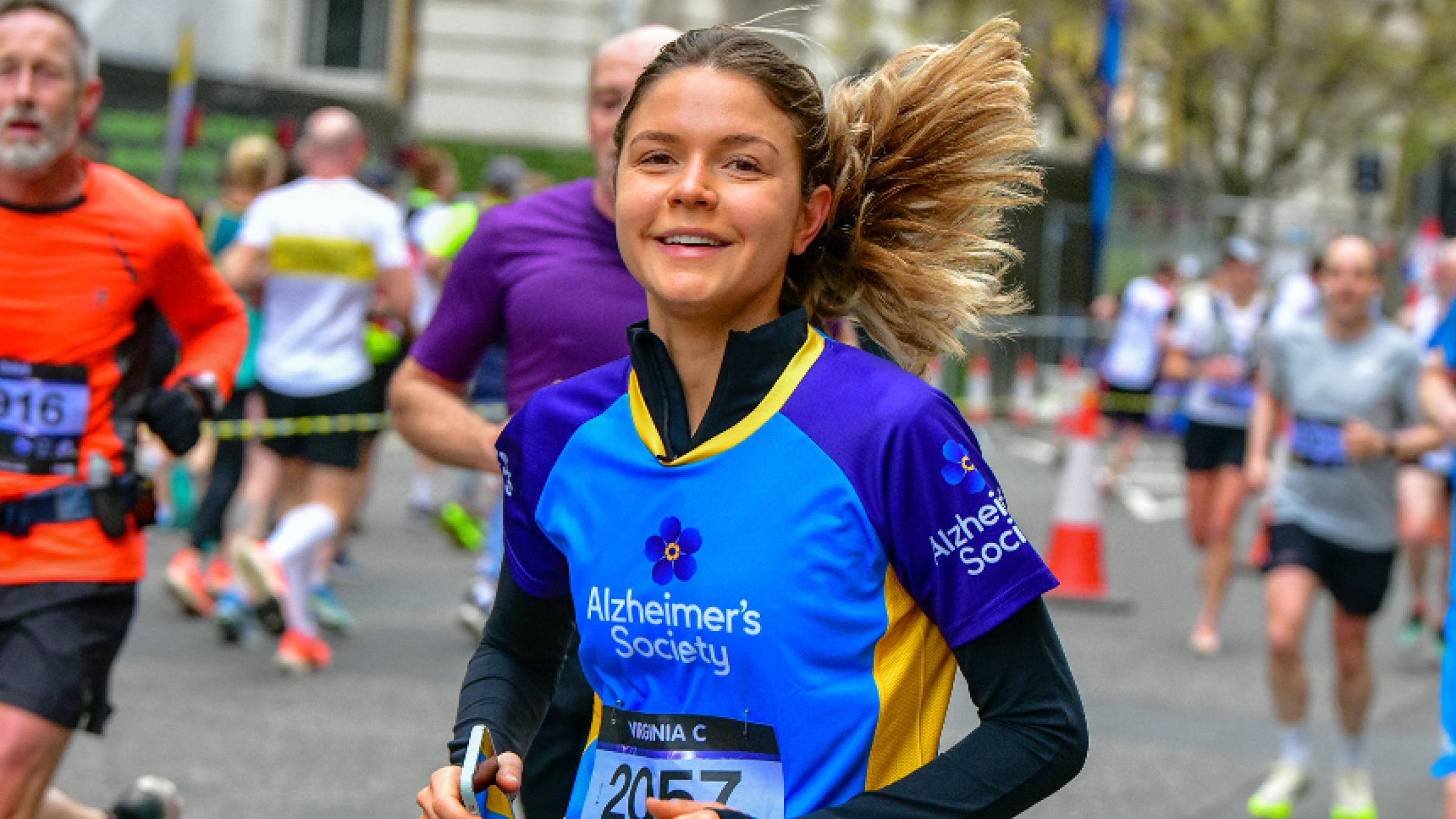 A group of runners through a city street with green trees in the background. A lady at the front of the group, in an Alzheimer's Society top, is smiling at the camera mid run.