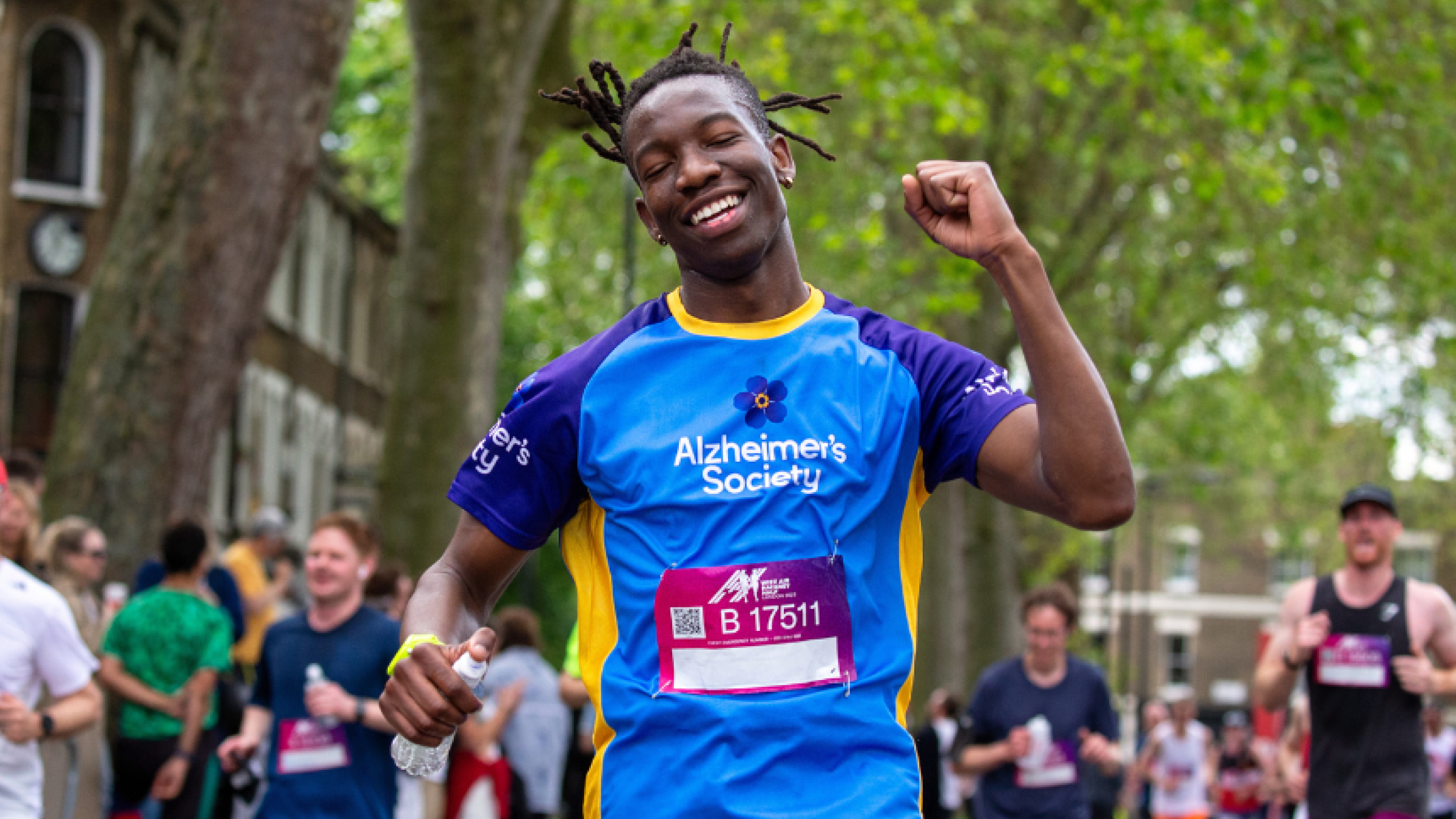 A gentleman punching the air with a mix of relief and tiredness on his face, with crowds around. He is dark skinned and wearing an Alzheimer's Society t-shirt and race number.