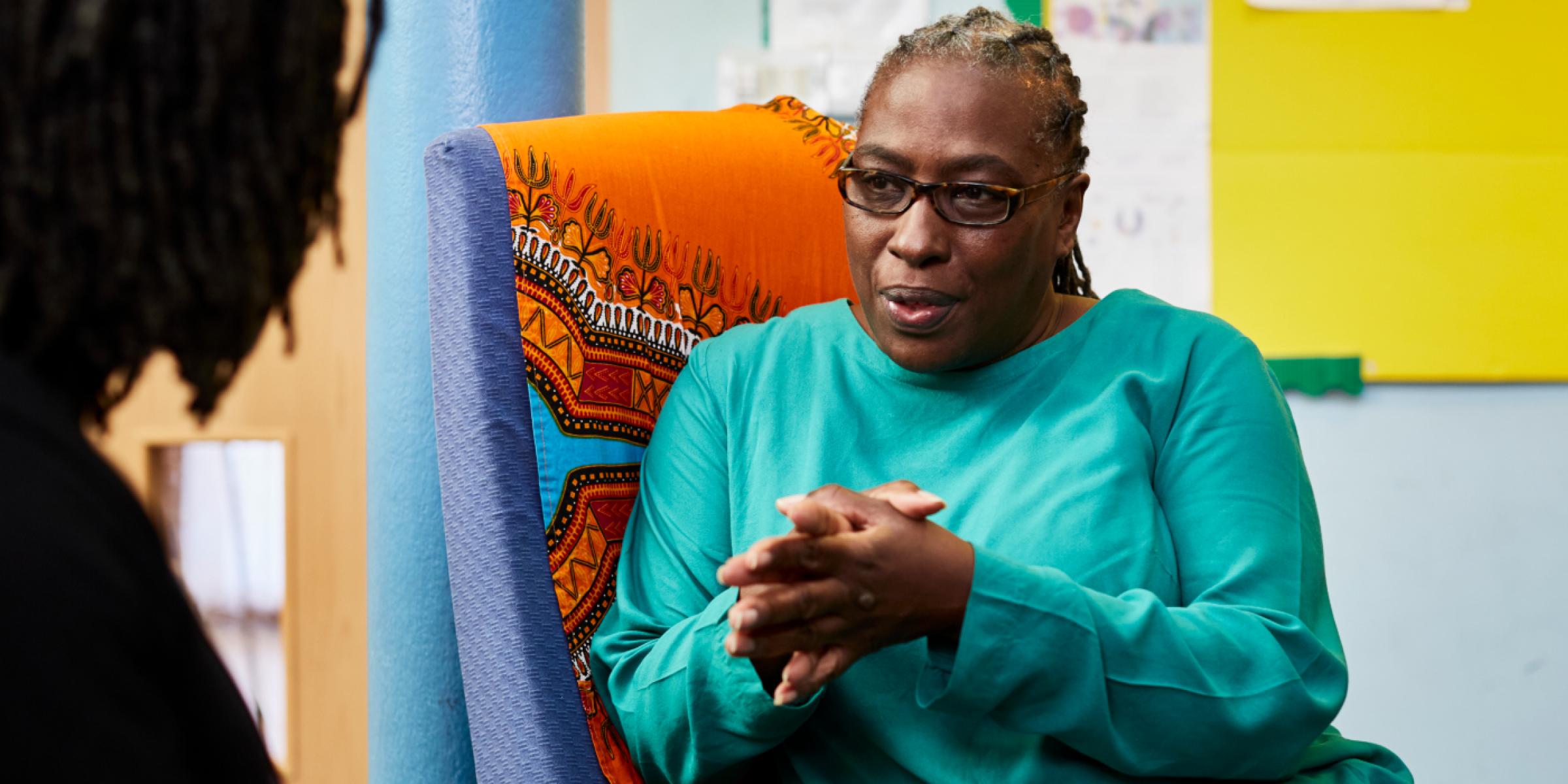 A Black woman in conversation with a person out of view, holding her hands together and listening intently