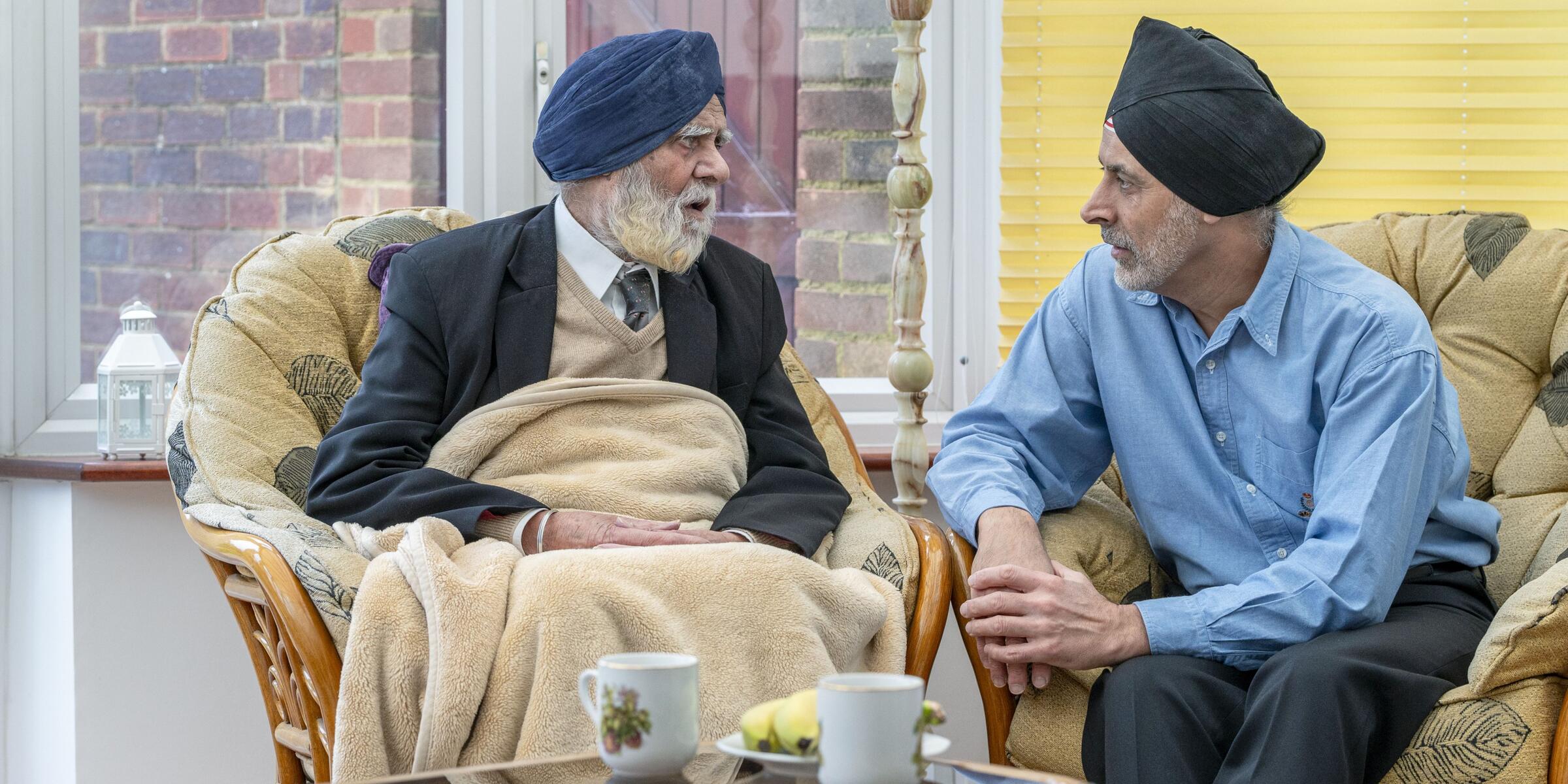 Two men sat on chairs. The man on the left has a blue turban, white beard, white shirt, beige vest, blue tie and jacket covered by a beige blanket and the men on the right wearing a blue turban, shirt and trousers