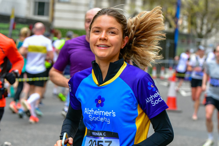 A group of runners through a city street with green trees in the background. A lady at the front of the group, in an Alzheimer's Society top, is smiling at the camera mid run.