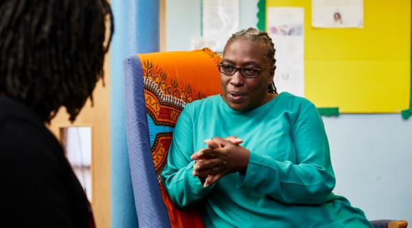 A Black woman in conversation with a person out of view, with her hands together and listening intently