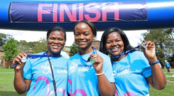 Three people wearing Memory Walk T-shirts holding their medals and smiling beneath a large inflatable 'Finish' line