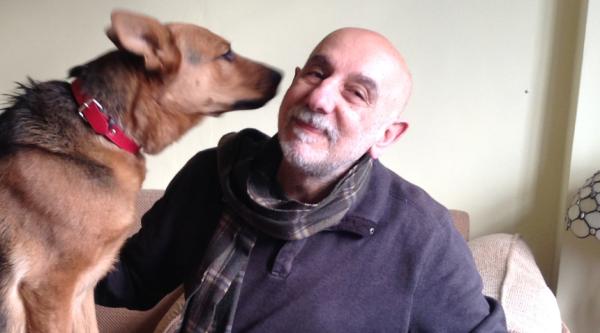 Armen sits on a sofa with a large brown dog holding its nose close to Armen's cheek, who is smiling