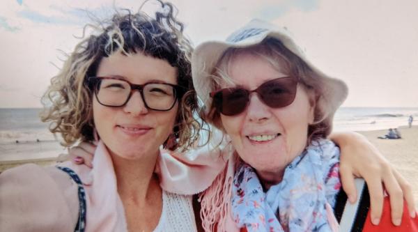Cara has her arm around her mum while standing on a sandy beach and smiling together