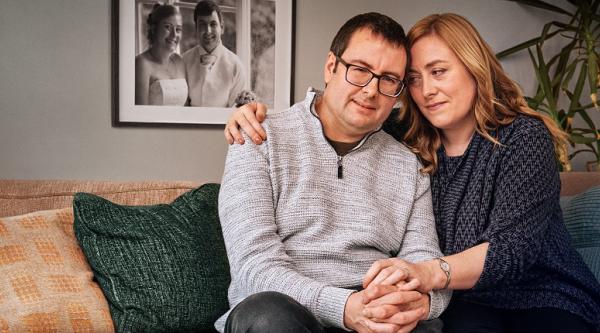 Mark and Caroline sit together on a sofa while holding hands