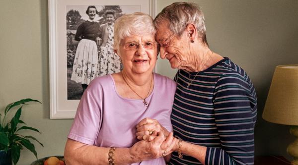 Hilary and Pauline in their living room. Hilary rests her head on Pauline's shoulder and holds her hand.
