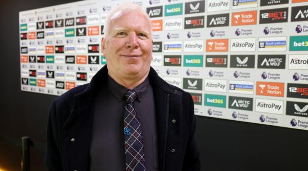 Peter Jones stood in front of a wall with advertisements on it at a football stadium, wearing an Alzheimer's Society pin badge