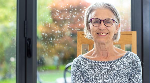 Anne Hoad sits in her home, her back to the door to her patio and garden. She is looking at the camera smiling and has short, grey hair. She wears glasses and a blue/ grey blouse