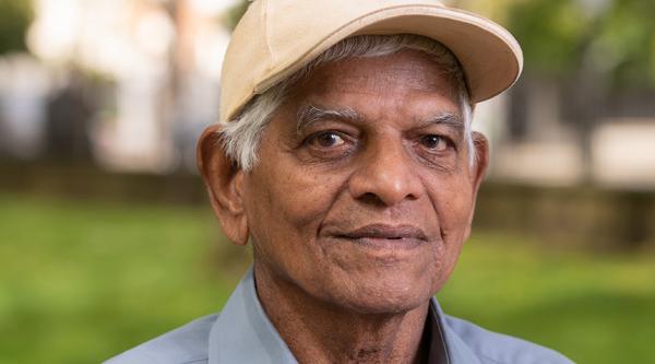 Lal stands outside in front of a green space. He is looking at the camera with a positive expression, wearing a beige baseball cap and light blue shirt