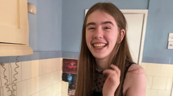 Eden has long brown hair and is wearing a black vest top. She's inside and smiling at the camera