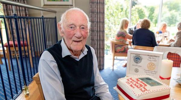 Billy is sat down wearing a navy tank top and blue shirt, next to him is a table with a celebration cake