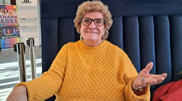 Maureen is wearing a bright yellow jumper and sat on a blue seating