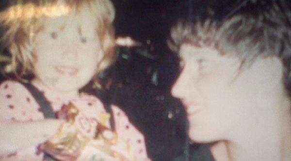 A toddler with ginger hair and a pink top eating crisps next to her smiling mum with dark brown hair