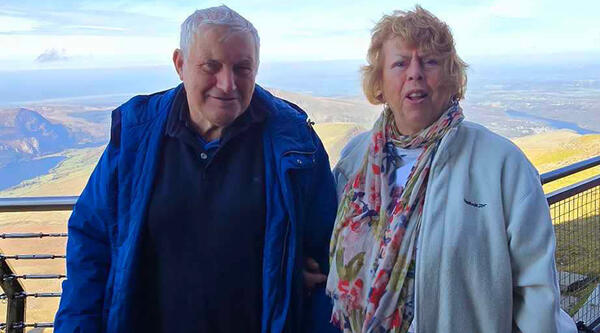 Man with white hair, wearing blue jacket and woman with blond hair, wearing white jacket and flowery scarf, with mountains behind them.