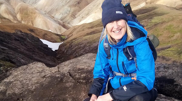 Woman with blond hair sat on rock in with mountains behind her wearing blue waterproof jacket and hat