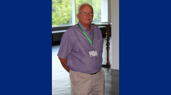 Man with white hair, wearing blue checked shirt and green lanyard in front of a window.