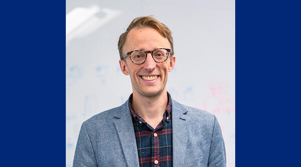 Man with brown hair and glasses, he is smiling and wearing a blue jacket and checked blue and red shirt