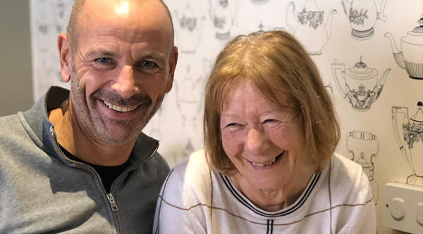 Man wearing grey top smiling, next to older woman who has mousy hair and is wearing a white top, there is patterned wallpaper behind them.