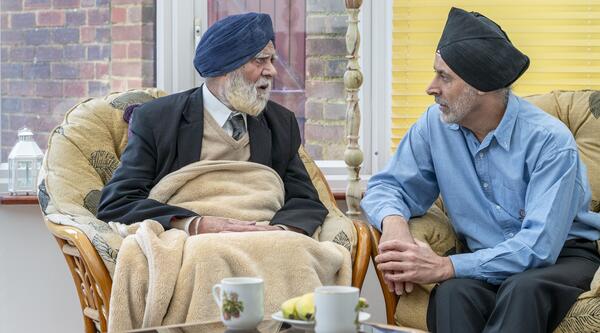 Two men sat on chairs. The man on the left has a blue turban, white beard, white shirt, beige vest, blue tie and jacket covered by a beige blanket and the men on the right wearing a blue turban, shirt and trousers
