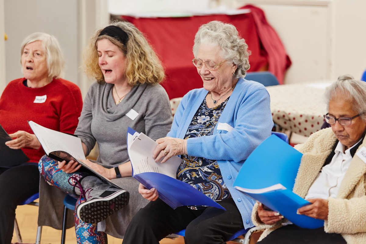 Four people sat in chairs looking at lyric sheets and singing