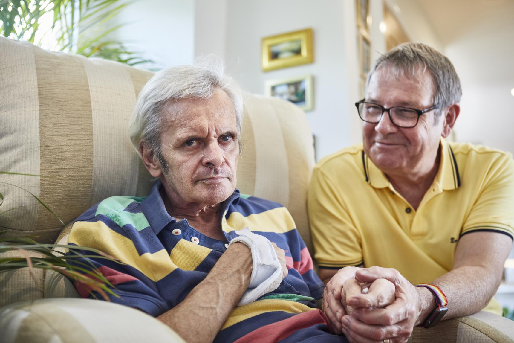 A man in a yellow top holds the hand of a man sat in a beige sofa wearing a colourful stripy top