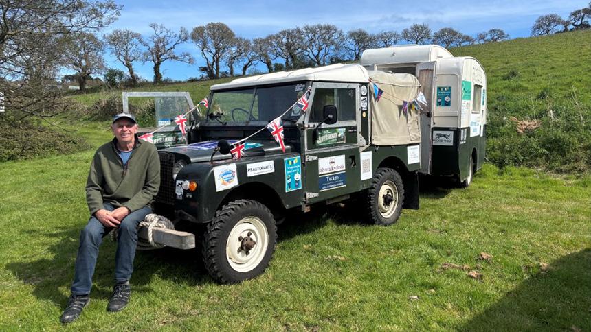 Derek sat on the bumper of his vintage Land Rover, which is parked in a field and pulling a vintage caravan