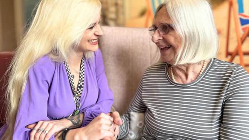 Kerri is talking to a care home resident and holding her hand. She's wearing a purple blouse and has long blonde hair
