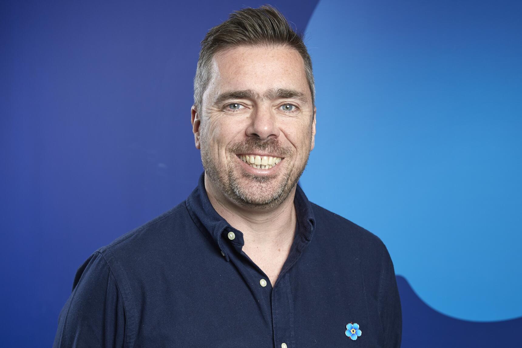 A man with short dark hair and a beard, wearing a blue shirt and smiling