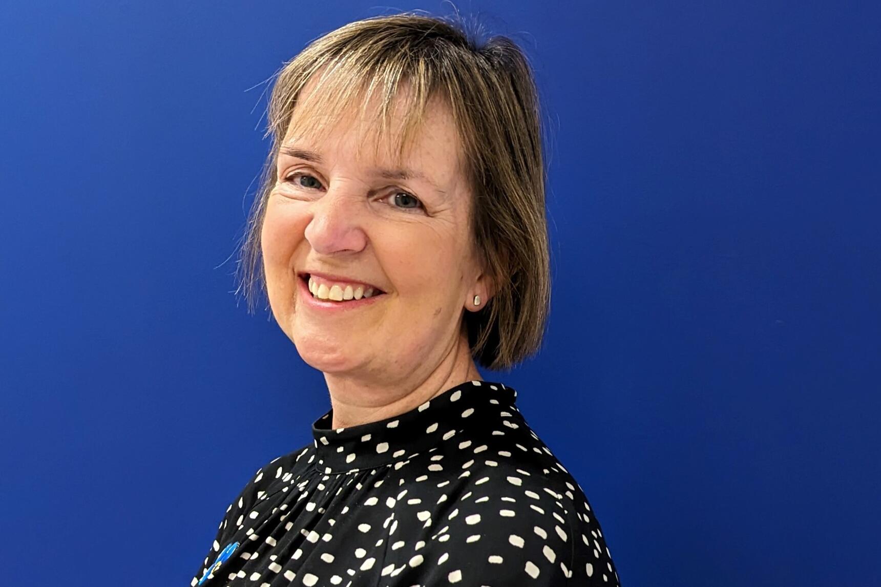 A lady with short blonde hair, smiling and wearing a black top with white dots