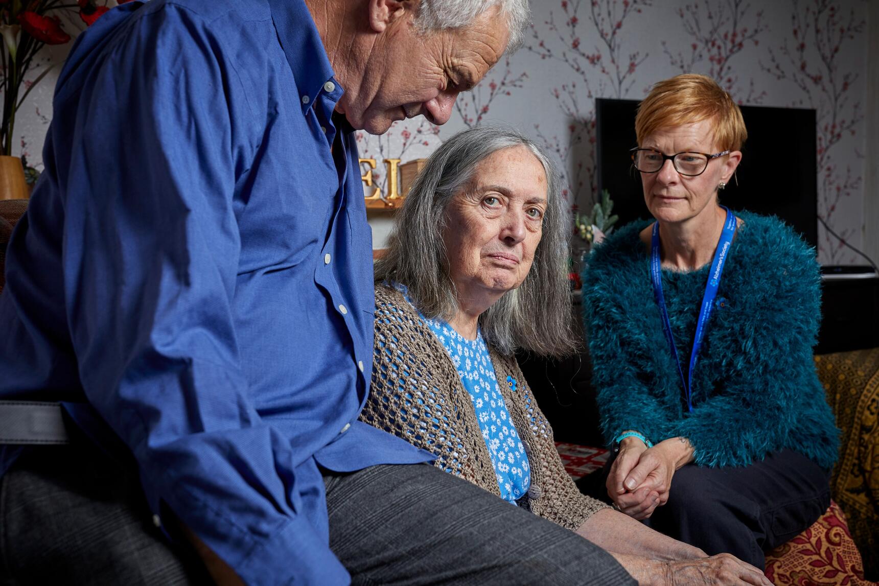 A woman with a grey bob looks at the camera, with her husband on the left and a support worker on the right