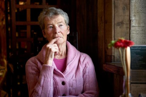 An older woman sits beside a window in a dimly lit room, deep in thought
