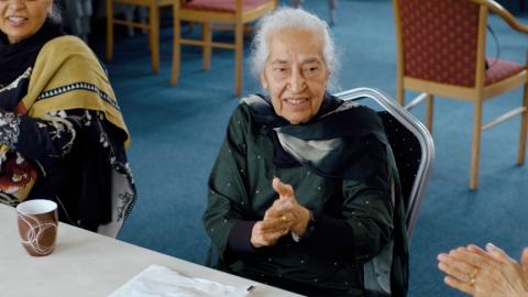 Bhagwant in her ladies group, sat in a chair smiling and clapping 