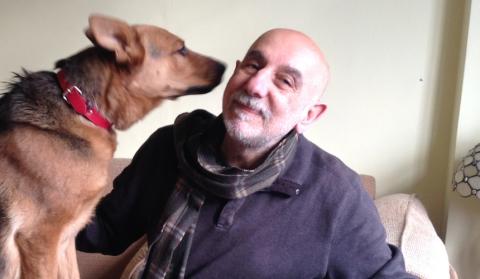 Armen sits on his sofa smiling while an old German Shepherd holds its nose close to Armen's right cheek