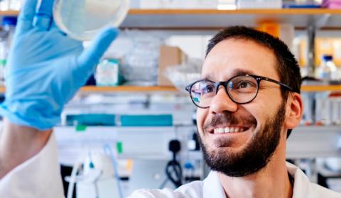 A researcher holding up a petri dish and smiling in a lab
