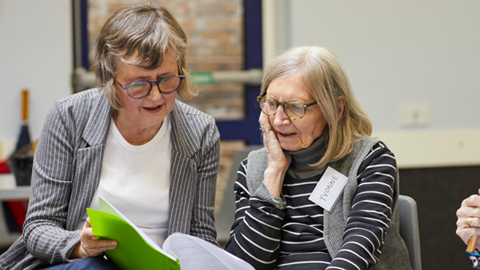 Two women sit side-by-side reading from sheets of paper.