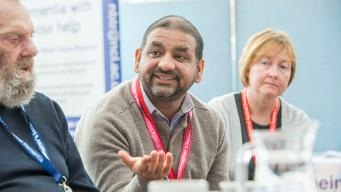 A man wearing a lanyard talks to his colleagues in a meeting.