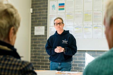 A woman wearing a dark blue Alzheimer's Society hoody stands at the front of a room speaking to a group