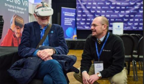 A man wears a VR headset while another man kneels next to him, both laughing.